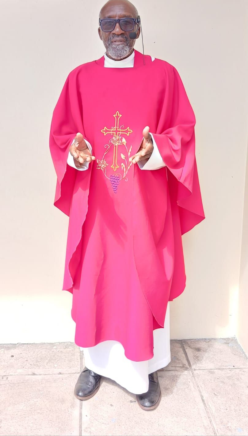 A person wearing a bright red chasuble over a white alb, featuring a gold cross emblem, standing with arms outstretched.