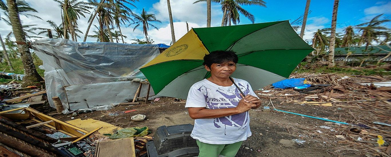 Woman holding a green and yellow umbrella stands near a makeshift shelter amidst debris. Palm trees and a cloudy sky are in the background.