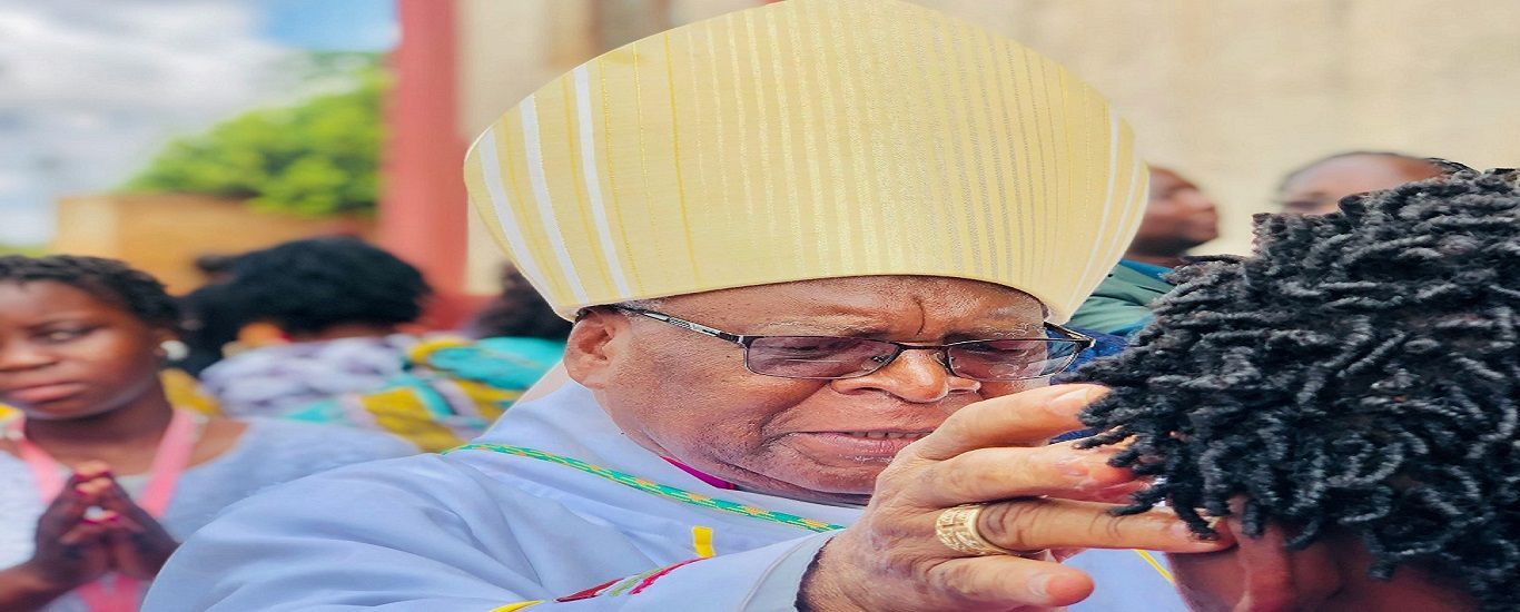 Bishop wearing a mitre, touching a person's head in an outdoor setting with other people present.