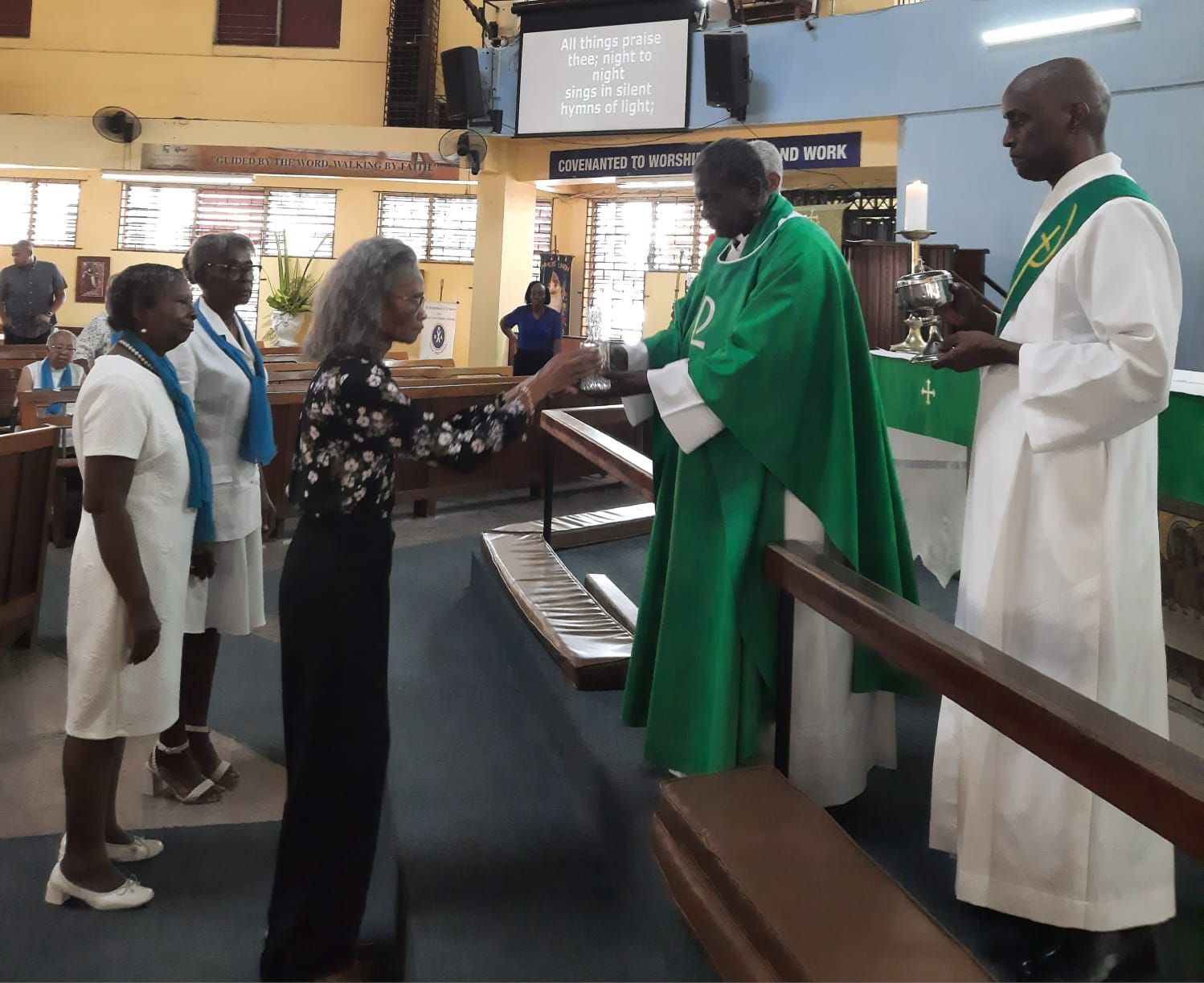 A priest in green robes offering communion to people in a church. Another man assists.