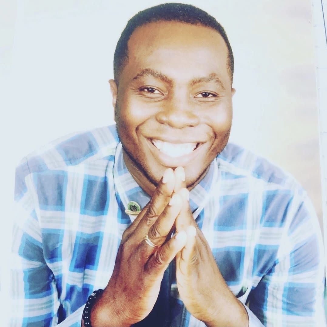 Man in blue plaid shirt smiling, hands clasped near face.