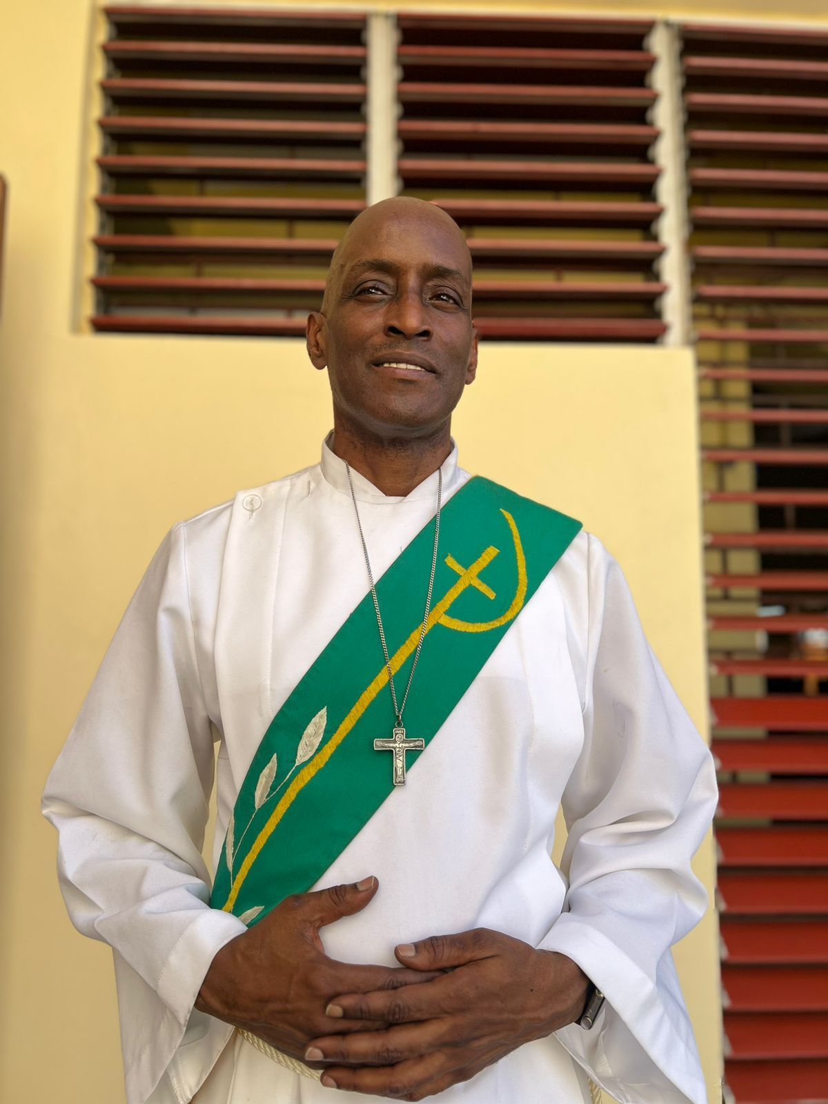 Clergyman with dark skin wearing white robe and green sash with cross necklace, standing indoors.