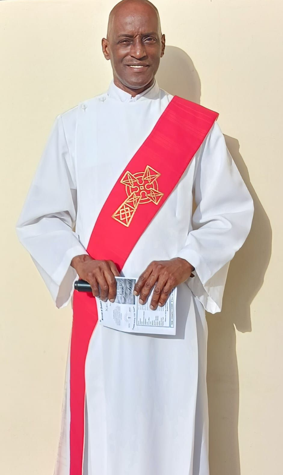 Clergyman with dark skin wearing white robe and green sash with cross necklace, standing indoors.