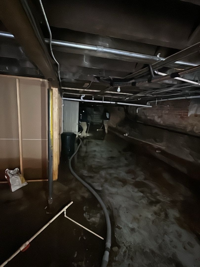 Dark, narrow crawl space with pipes, electrical wiring, and a hose. Wet flooring and exposed wooden beams.