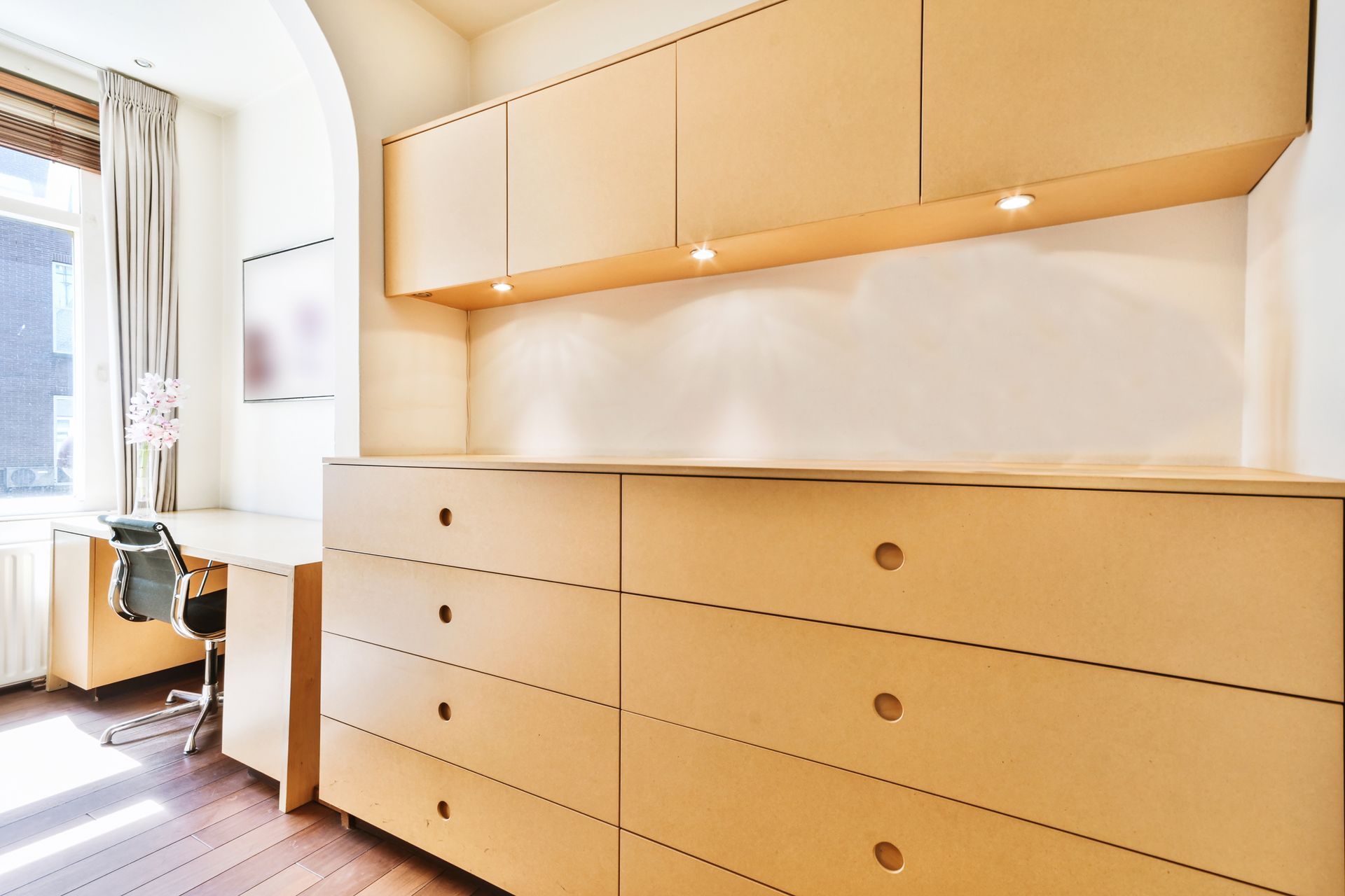 Built-in wooden cabinetry in a room with a desk and window. Cabinets are light brown, with recessed lighting.