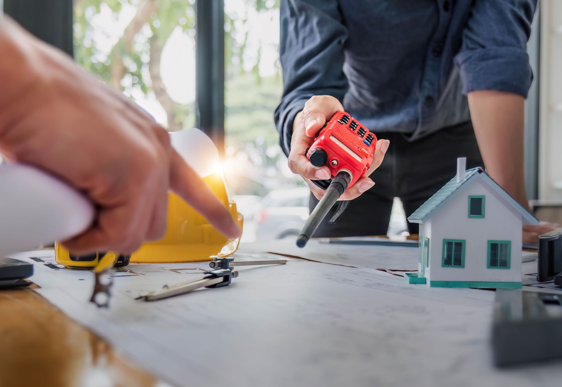 Two people reviewing blueprints, one holding a walkie-talkie and another pointing at a yellow hard hat and a model house on a desk.