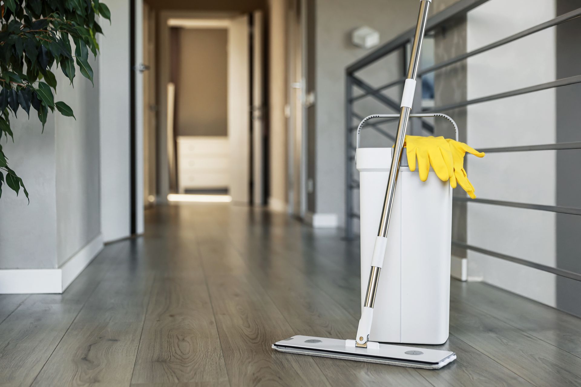 Mop and bucket with yellow gloves rest on a wooden floor in a hallway near a railing.