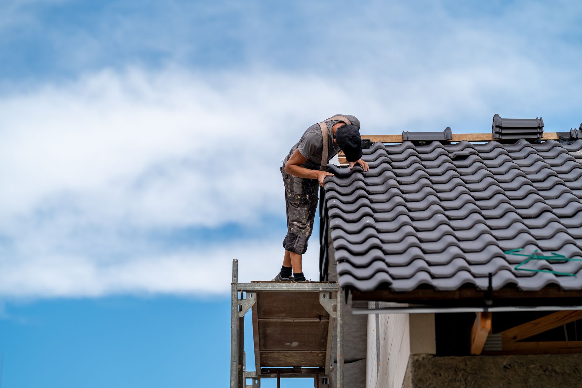 Roofer on scaffolding installing dark gray roof tiles against a blue sky.