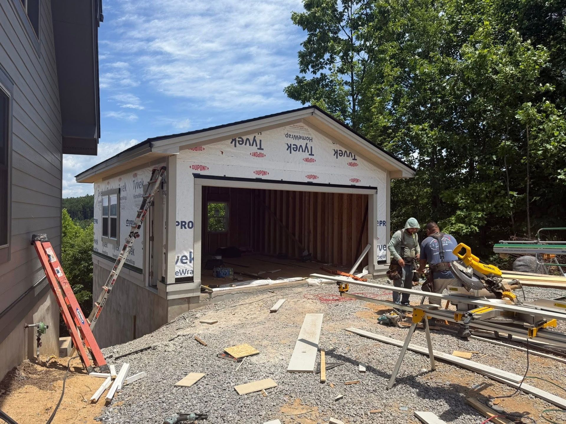 Garage under construction next to a house. Workers with tools, blue sky, and gravel ground.