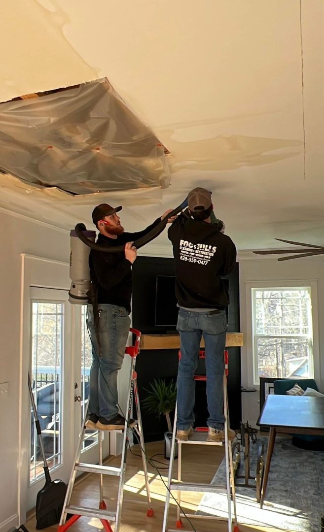 Two people vacuuming a ceiling, one on a ladder with a backpack vacuum, in a living room with plastic sheeting.