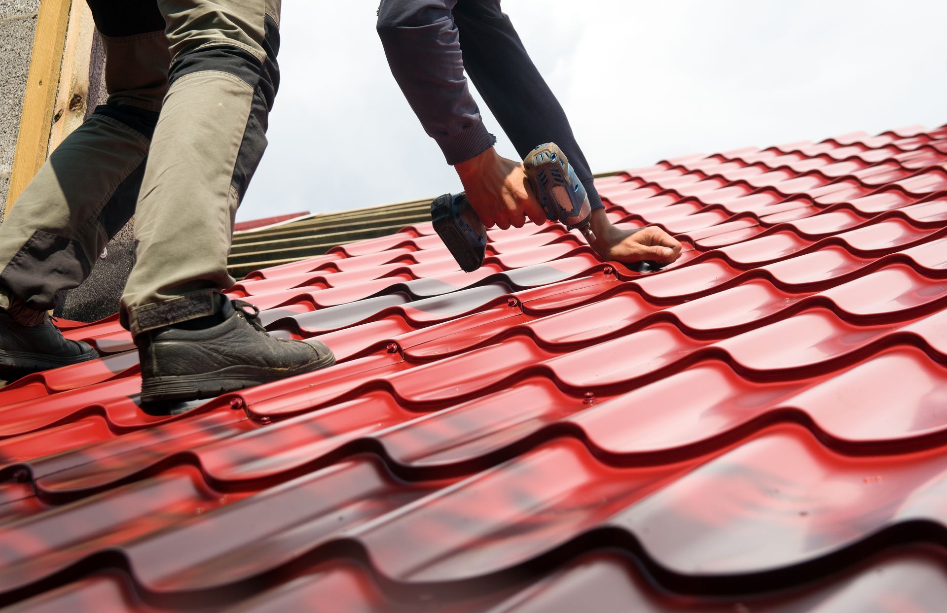Two workers installing a red metal roof using a power drill.