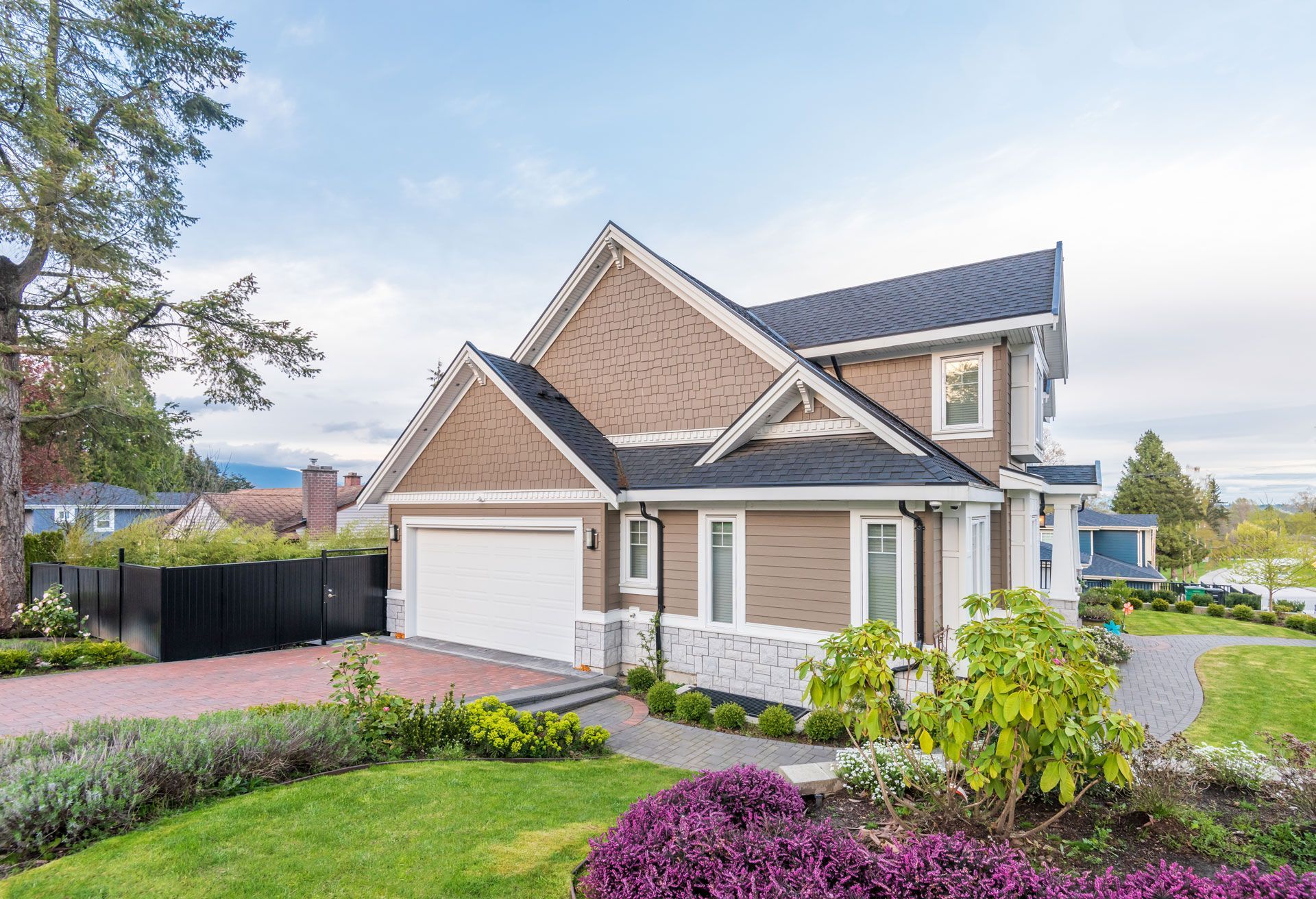Two-story brown house with a white garage door, surrounded by landscaping and a brick driveway.