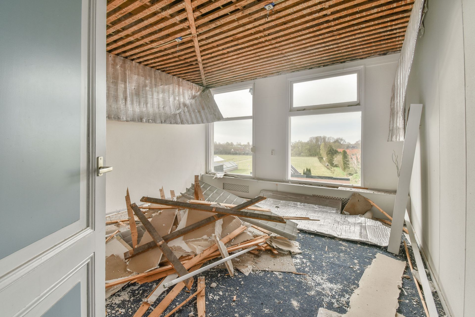 Room under construction, exposed beams, debris on floor, windows overlooking greenery, door on left.