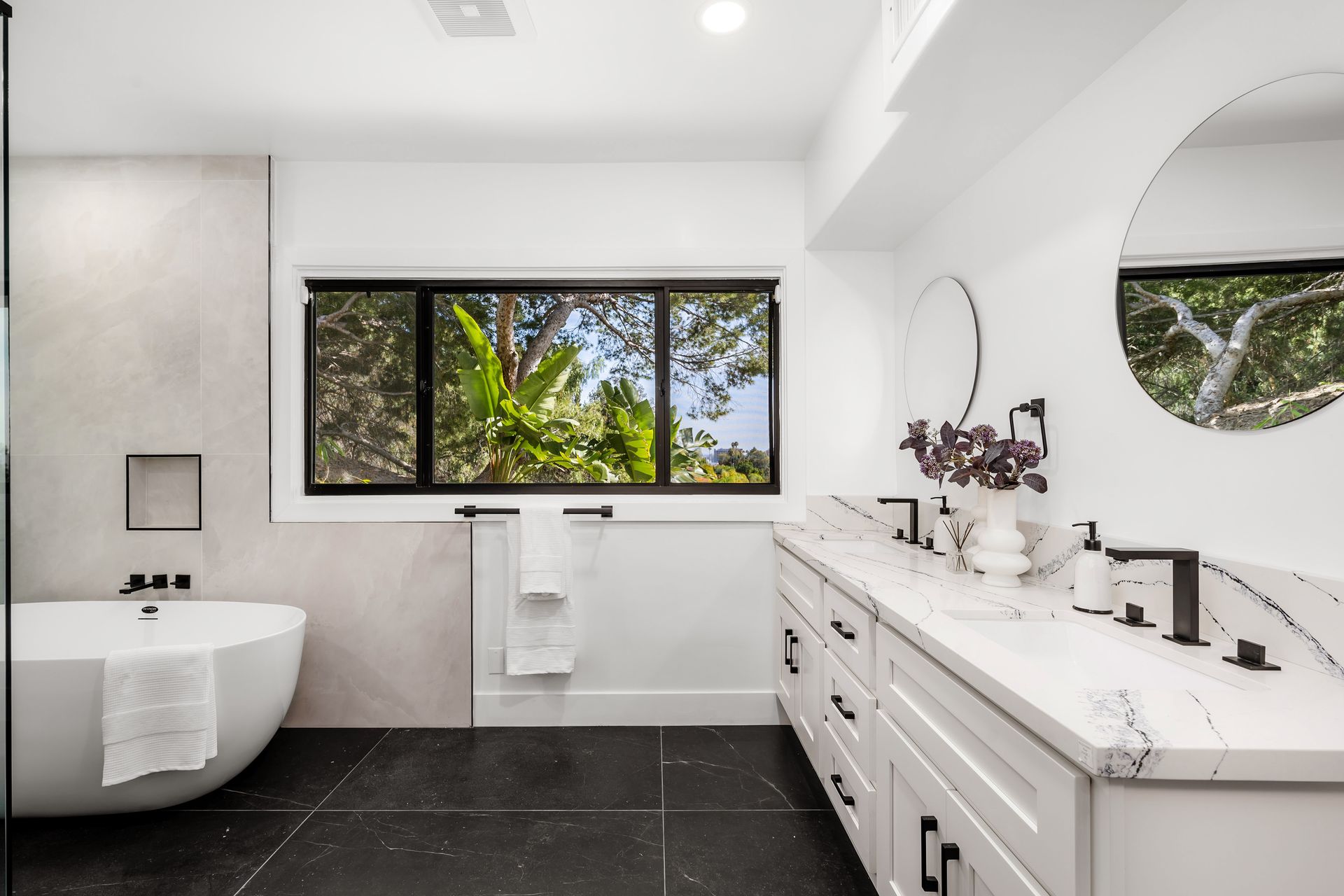 Modern bathroom with white cabinets, marble countertop, black trim window, and freestanding tub.