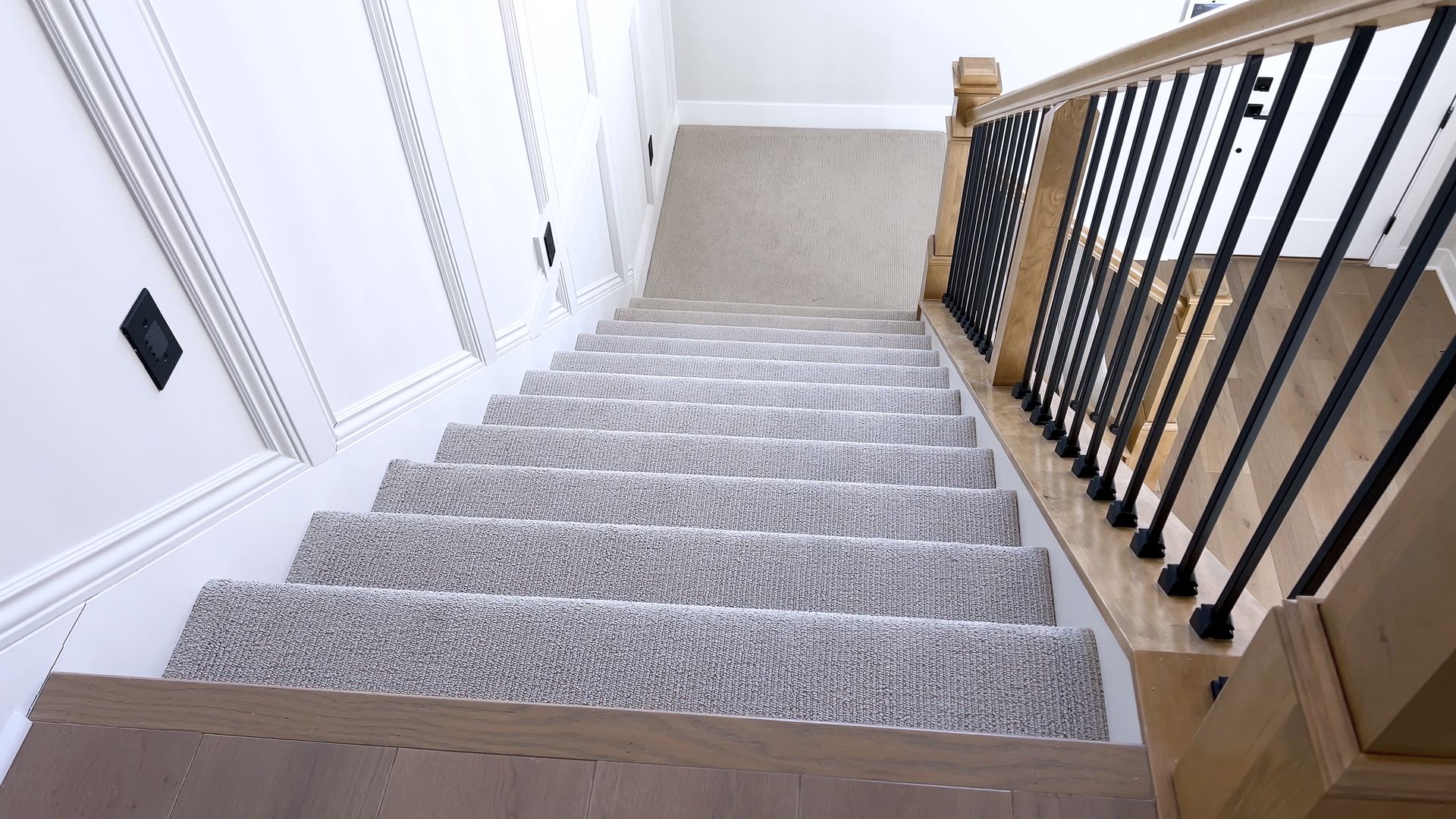 Staircase with carpeted steps, wooden handrail with black spindles, and white paneled walls.