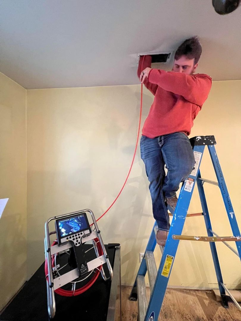 Person on ladder installs cable into ceiling, connected to a machine with a screen. Interior setting.