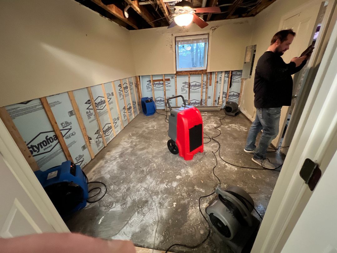 Man installing drywall in a room with exposed insulation. Dehumidifiers and air movers are present, indicating water damage.
