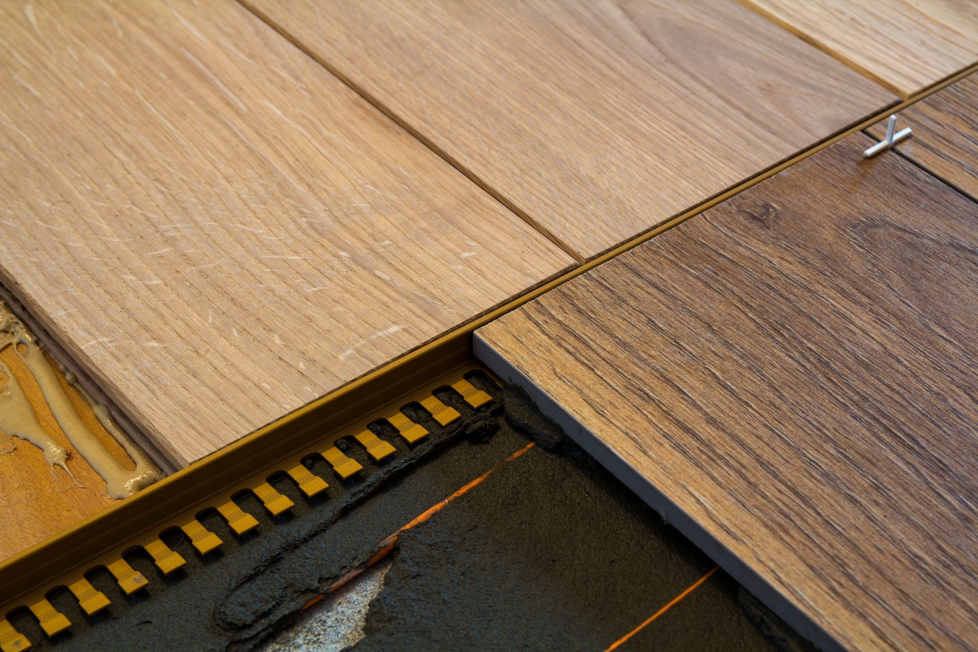 Close-up of hardwood flooring being installed, showing a metal edge piece, spacers, and mortar.