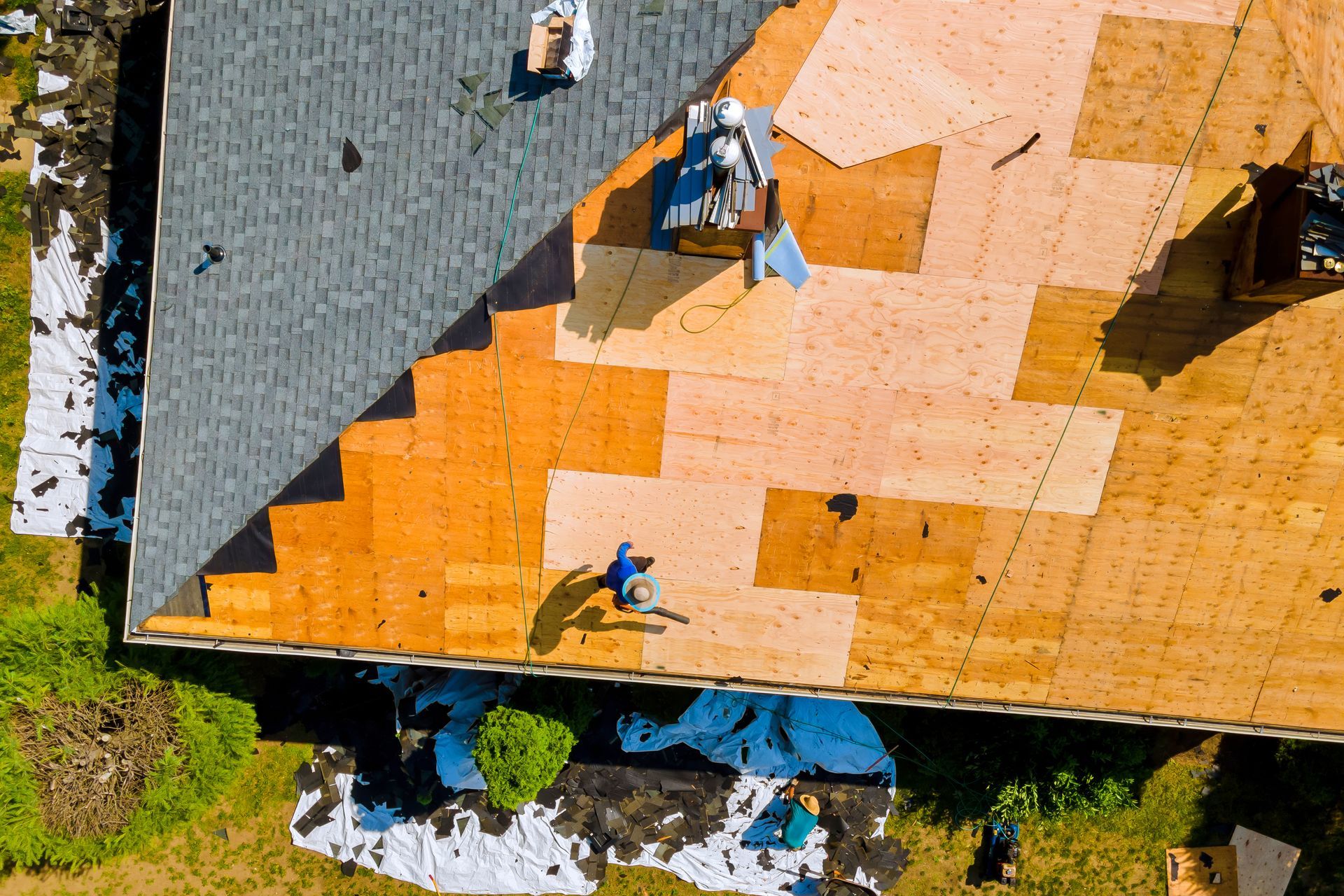 Roof being re-shingled; worker on partially exposed wood roof, laying down new shingles. Blue tarp, debris, and gray existing shingles visible.
