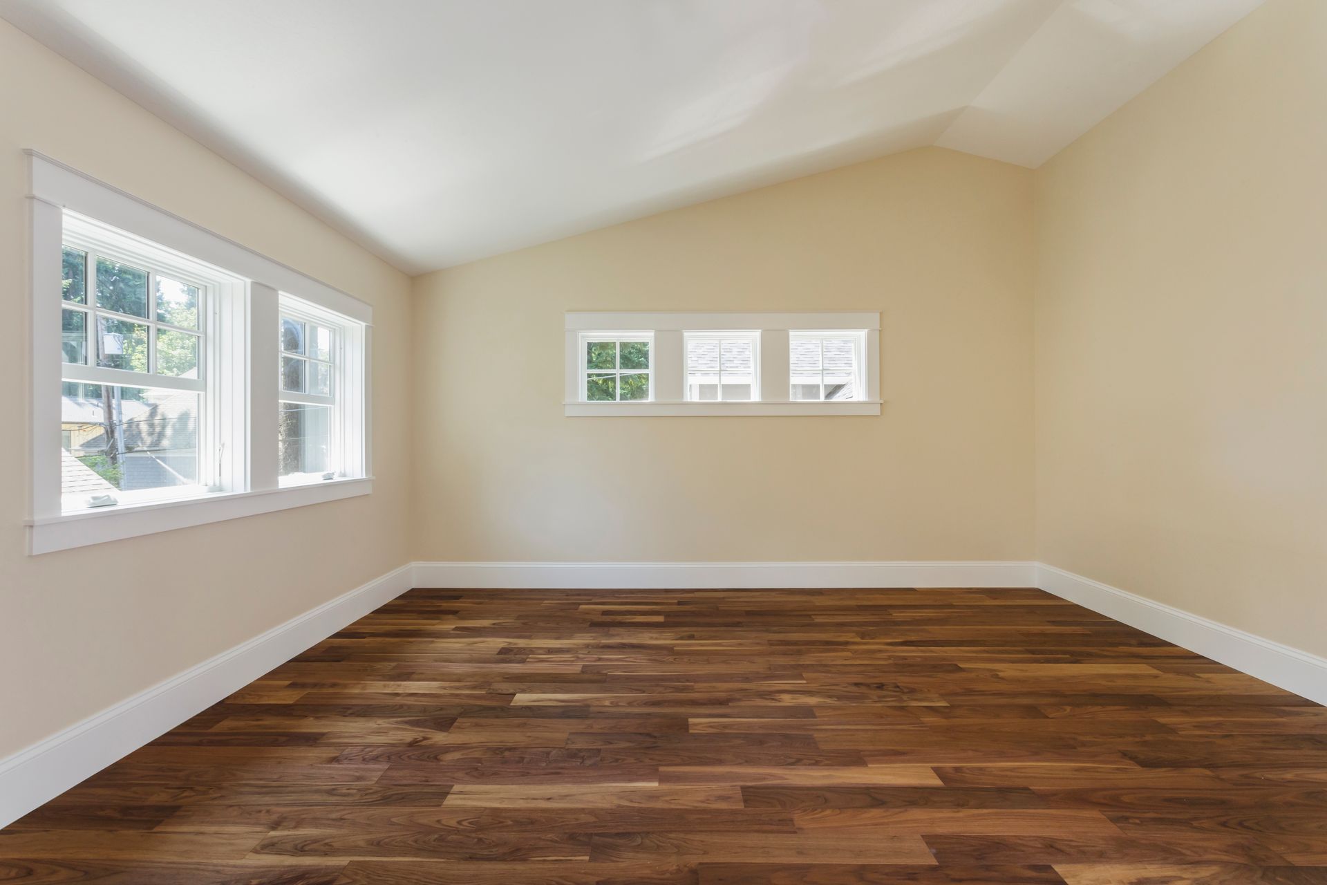 Empty room with hardwood floors, two windows on one wall, three smaller windows on another. Beige walls and white trim.