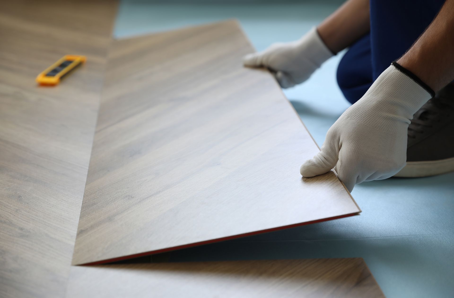 Person installing flooring, holding a plank. White gloves, light-colored wood-look plank, blue floor.
