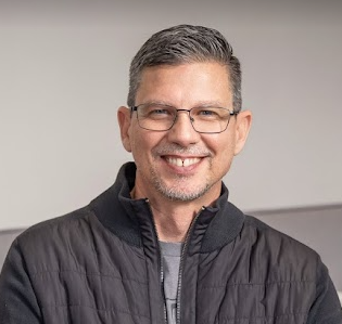 A man wearing glasses and a black jacket is smiling for the camera.
