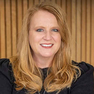 A woman with long blonde hair is smiling for the camera in front of a wooden wall.
