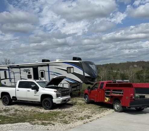 White pickup with open hood beside red utility truck and RV in a rocky field under cloudy sky