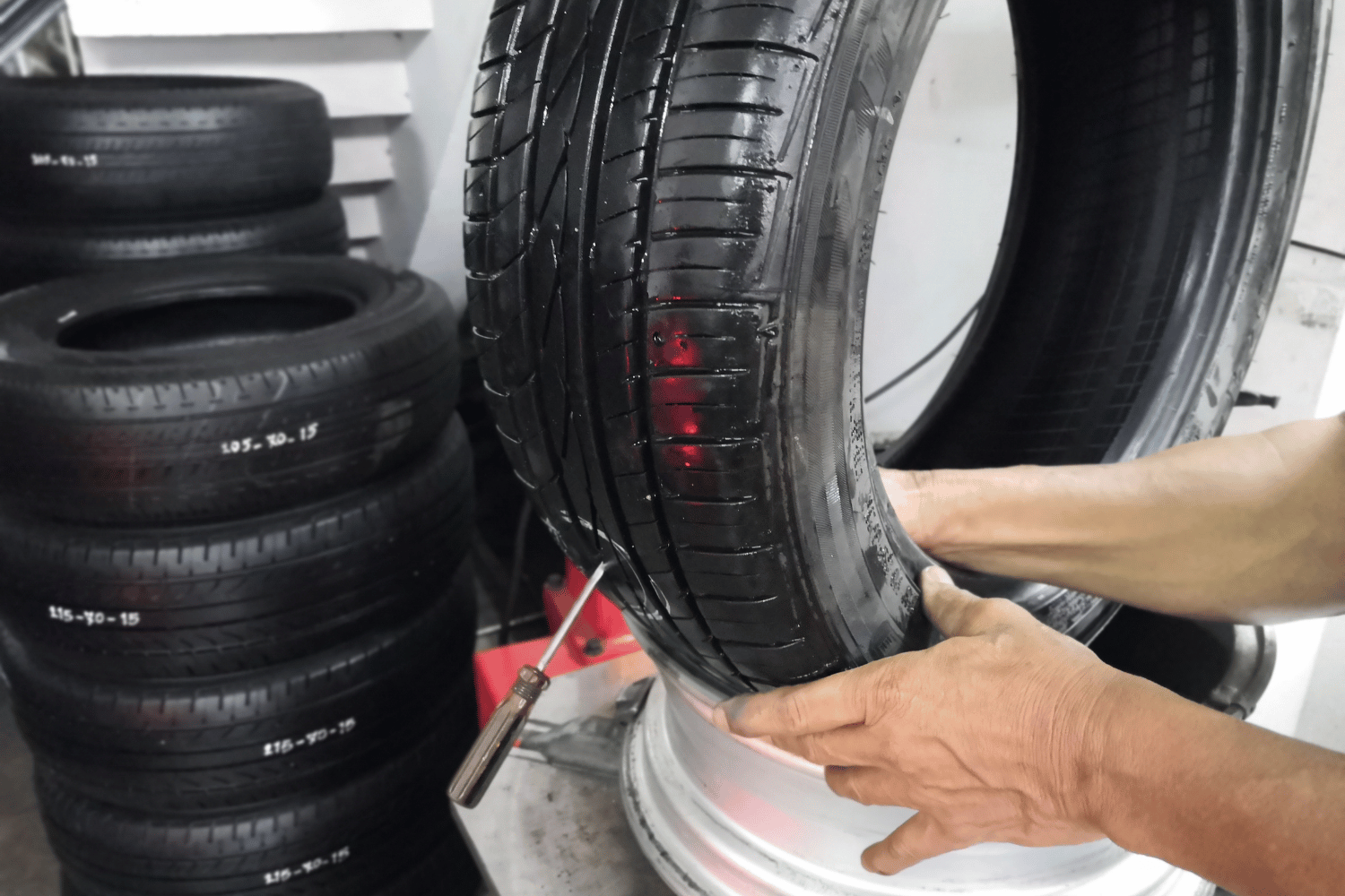 Hands mounting a car tire on a rim in a workshop, with stacked tires in the background
