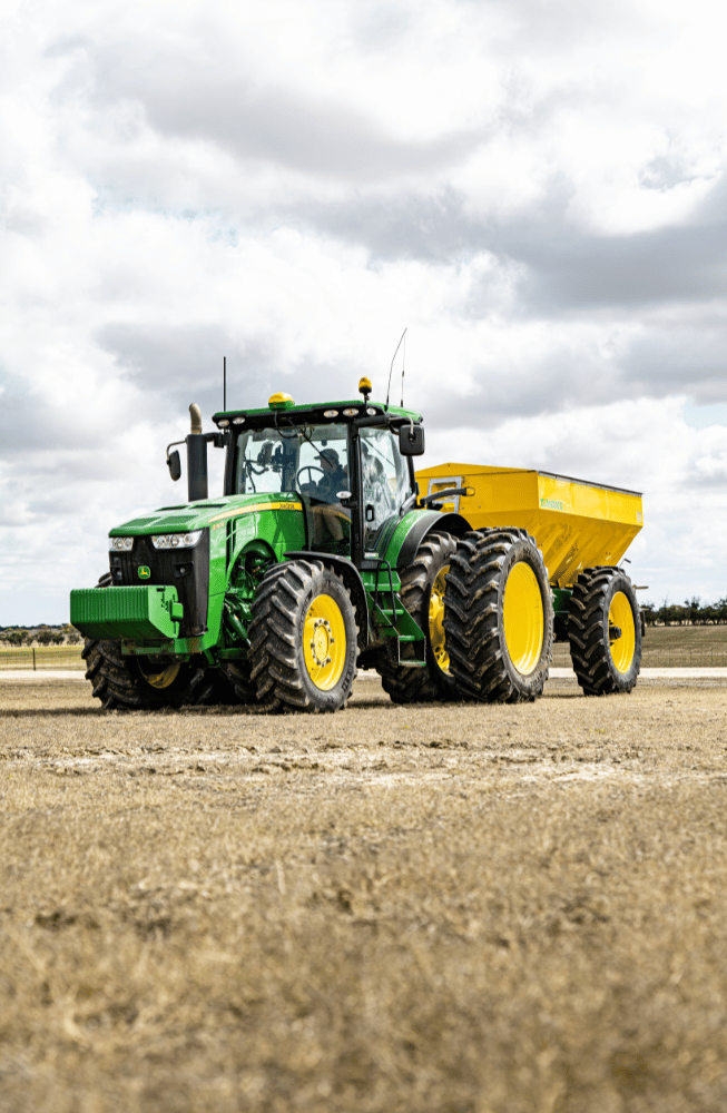 Green tractor hauling a yellow trailer across a dirt field under cloudy skies