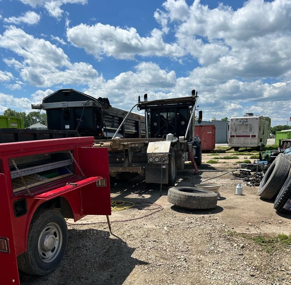 Damaged trucks and equipment in a gravel yard under a cloudy sky, with a red truck at left.