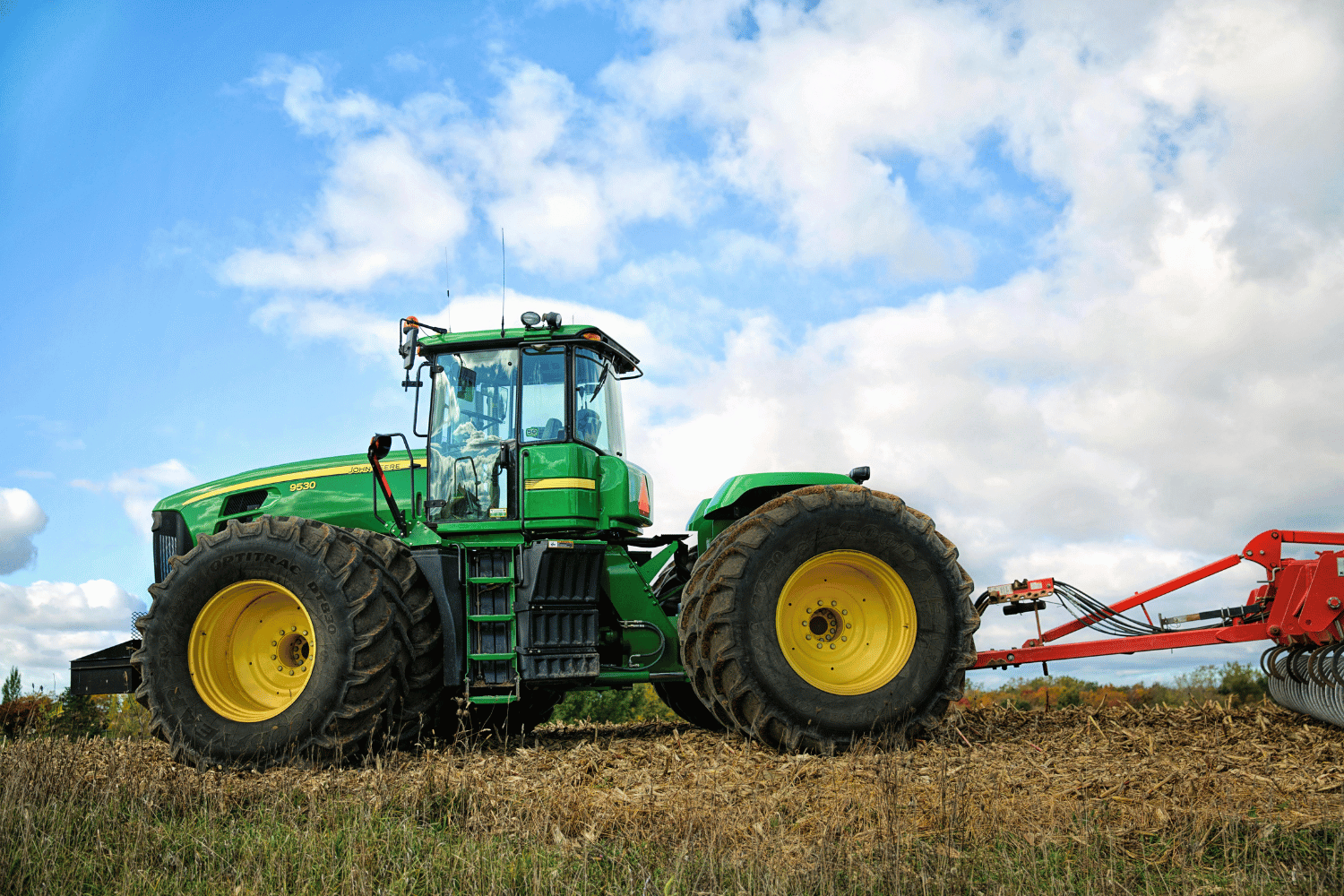 Green tractor in a field under a cloudy blue sky, towing red farm equipment.