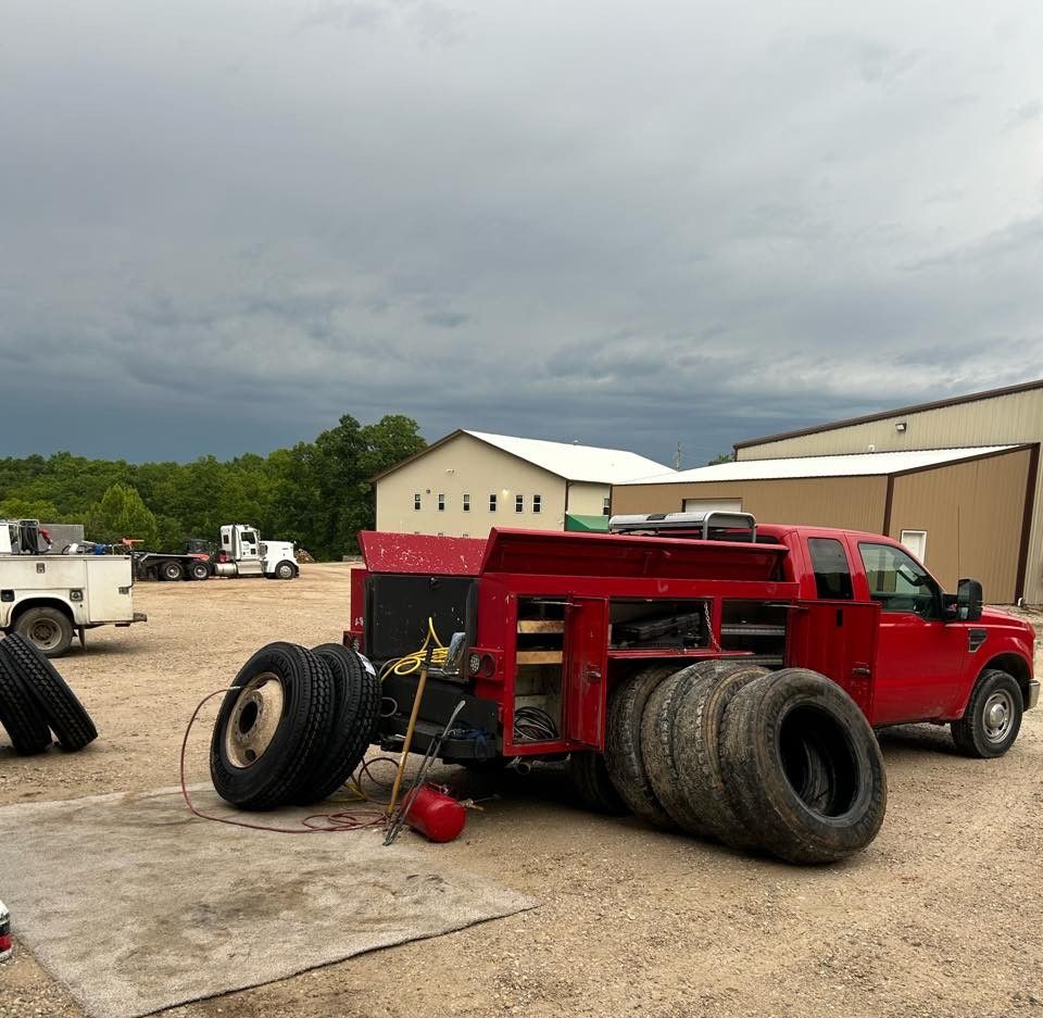 Red off-road truck with oversized rear tires parked on a gravel lot under cloudy skies