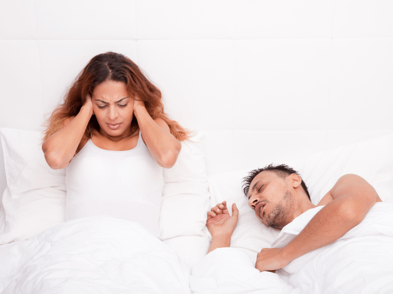 A woman is covering her ears while a man sleeps in bed snoring.