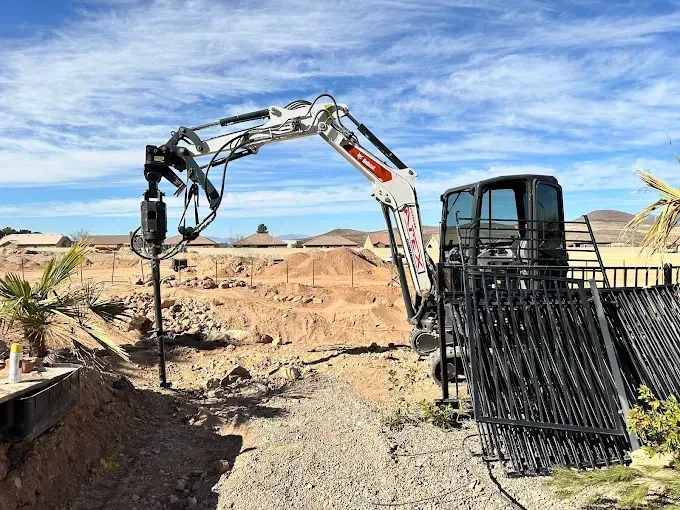 A Bobcat mini excavator drilling a hole for a fence post in a residential yard. Blue sky and dirt mound in background.