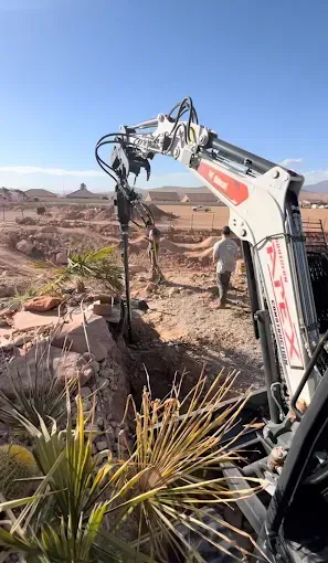 Bobcat excavator drilling in rocky terrain; person walks in background; clear blue sky.