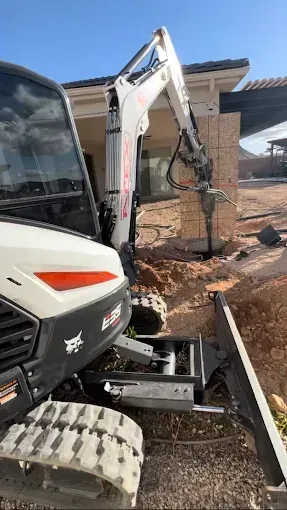A white Bobcat excavator uses a jackhammer to break up a brick structure near a house.