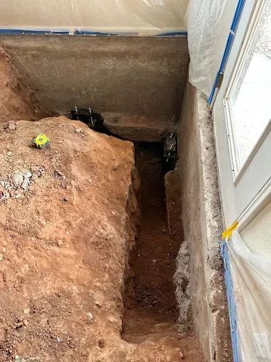 Trench dug near a building's entrance. Red soil, concrete foundation, white door. Yellow tape, plastic sheeting.