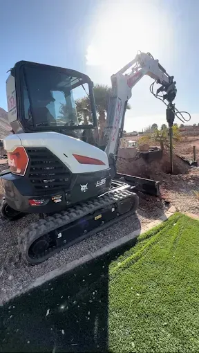 Bobcat excavator on a gravel base, drilling into soil under a sunny sky.