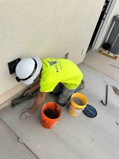 Person in hard hat patching concrete near a building. Buckets and tools are nearby.
