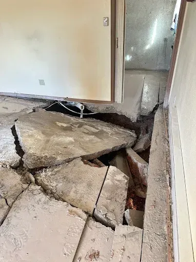 Collapsed floor in a room, revealing debris and structural damage. Doorway leads to another space.