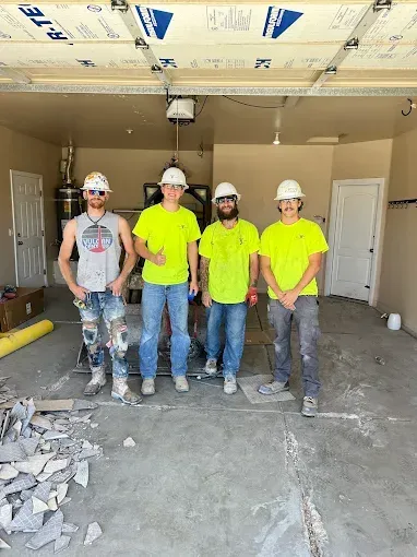 Four construction workers in a garage wearing hard hats and neon yellow shirts; debris on the floor.