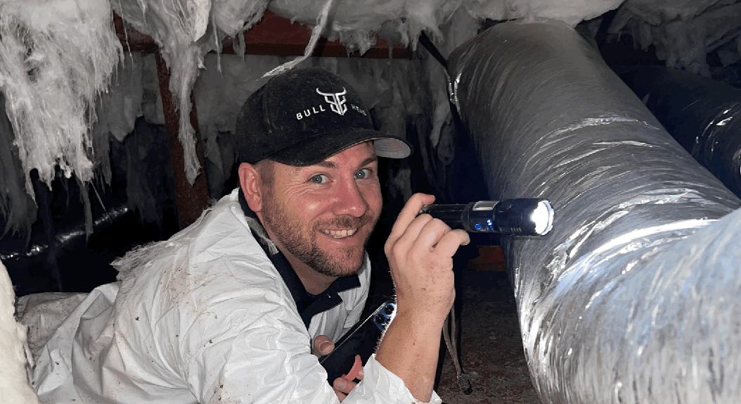 Man in white protective suit, holding a flashlight in a crawl space.