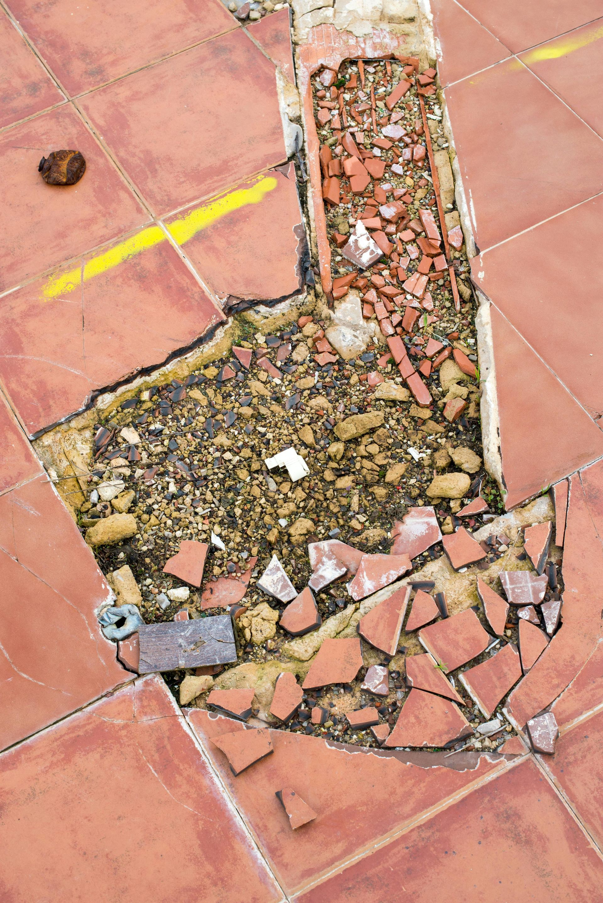 Damaged red tile floor with a large hole revealing gravel and debris. Yellow marking line.