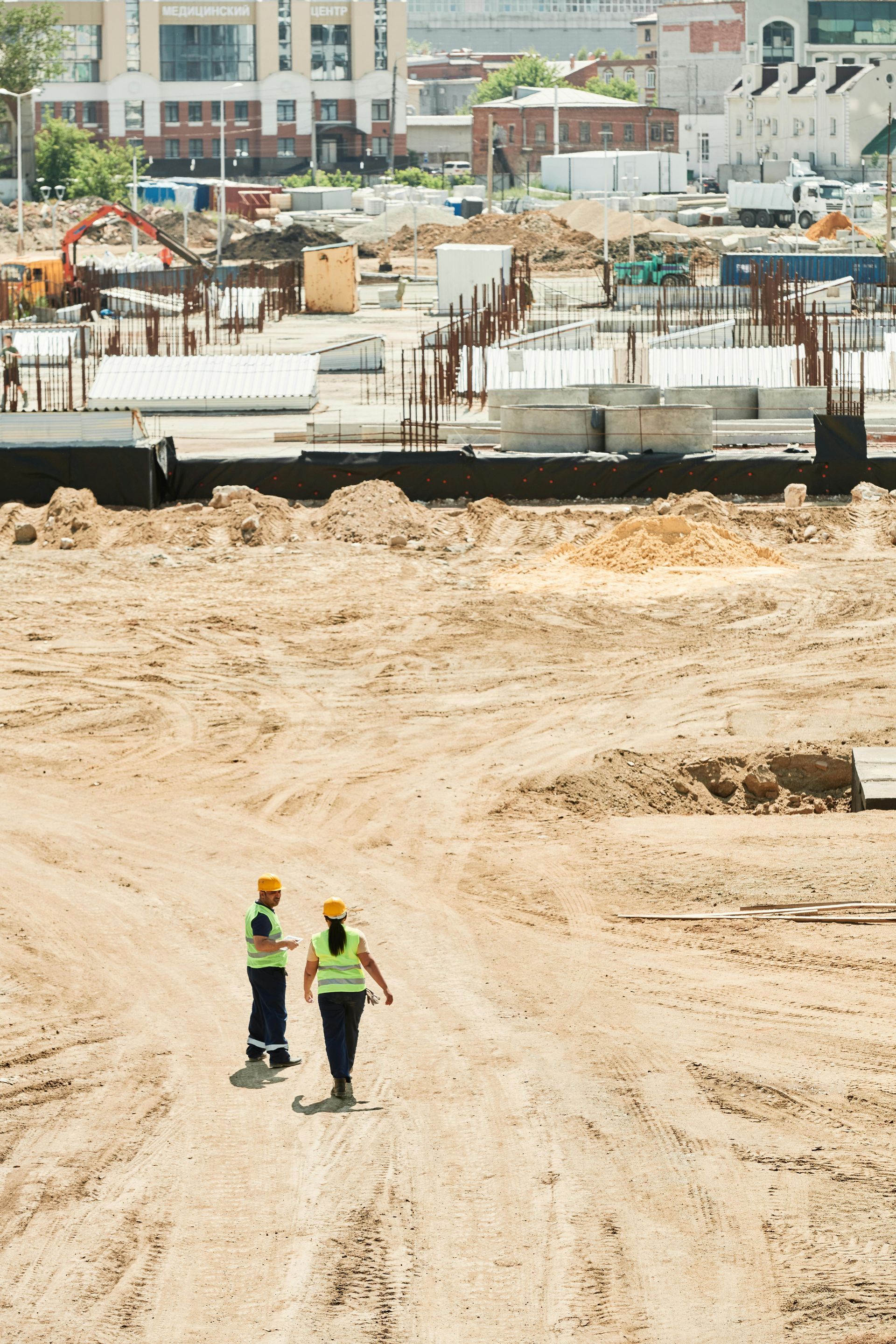 Two construction workers in safety vests walk on a dusty construction site.