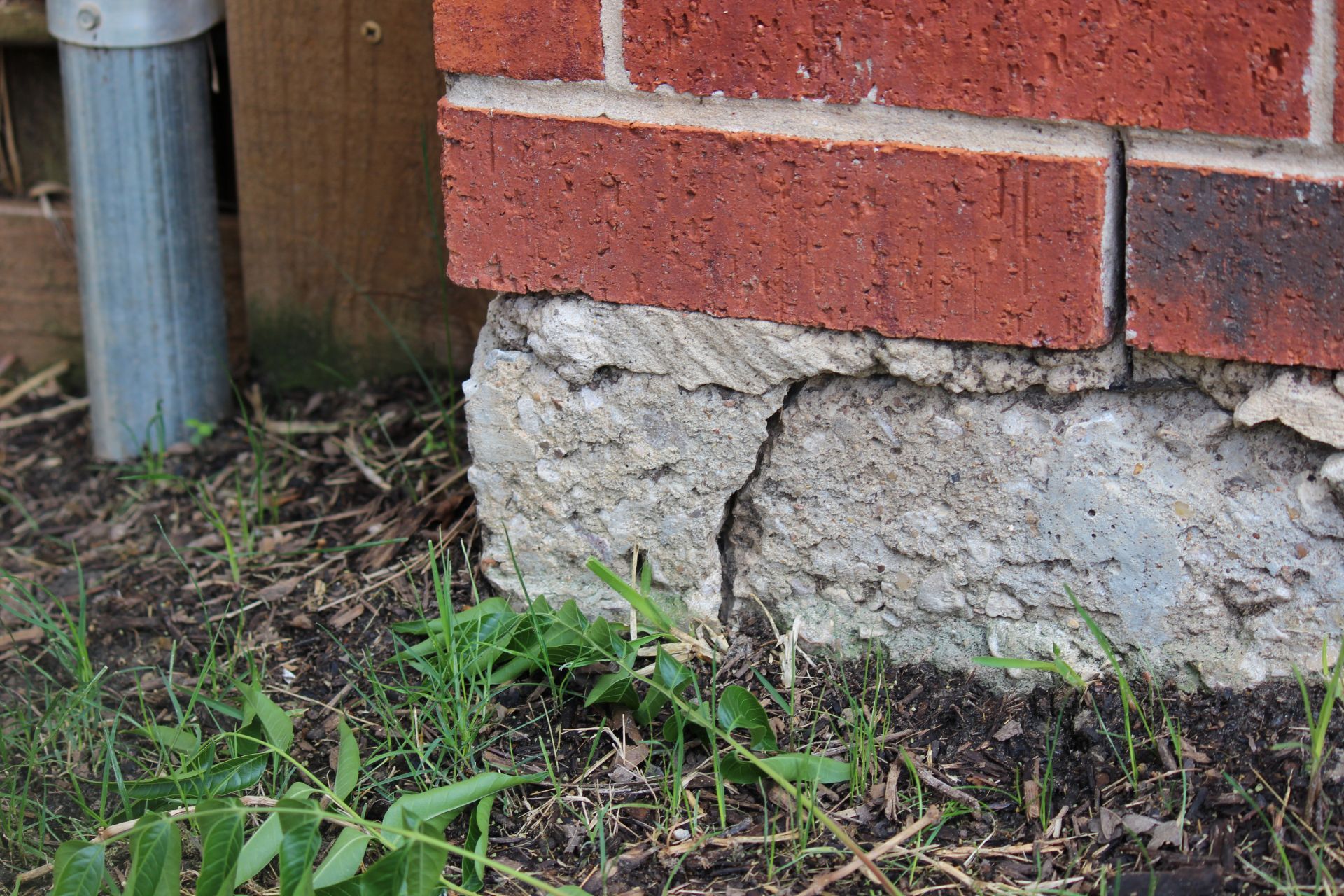 Brick wall section resting on a cracked concrete foundation near grass and a metal pipe.