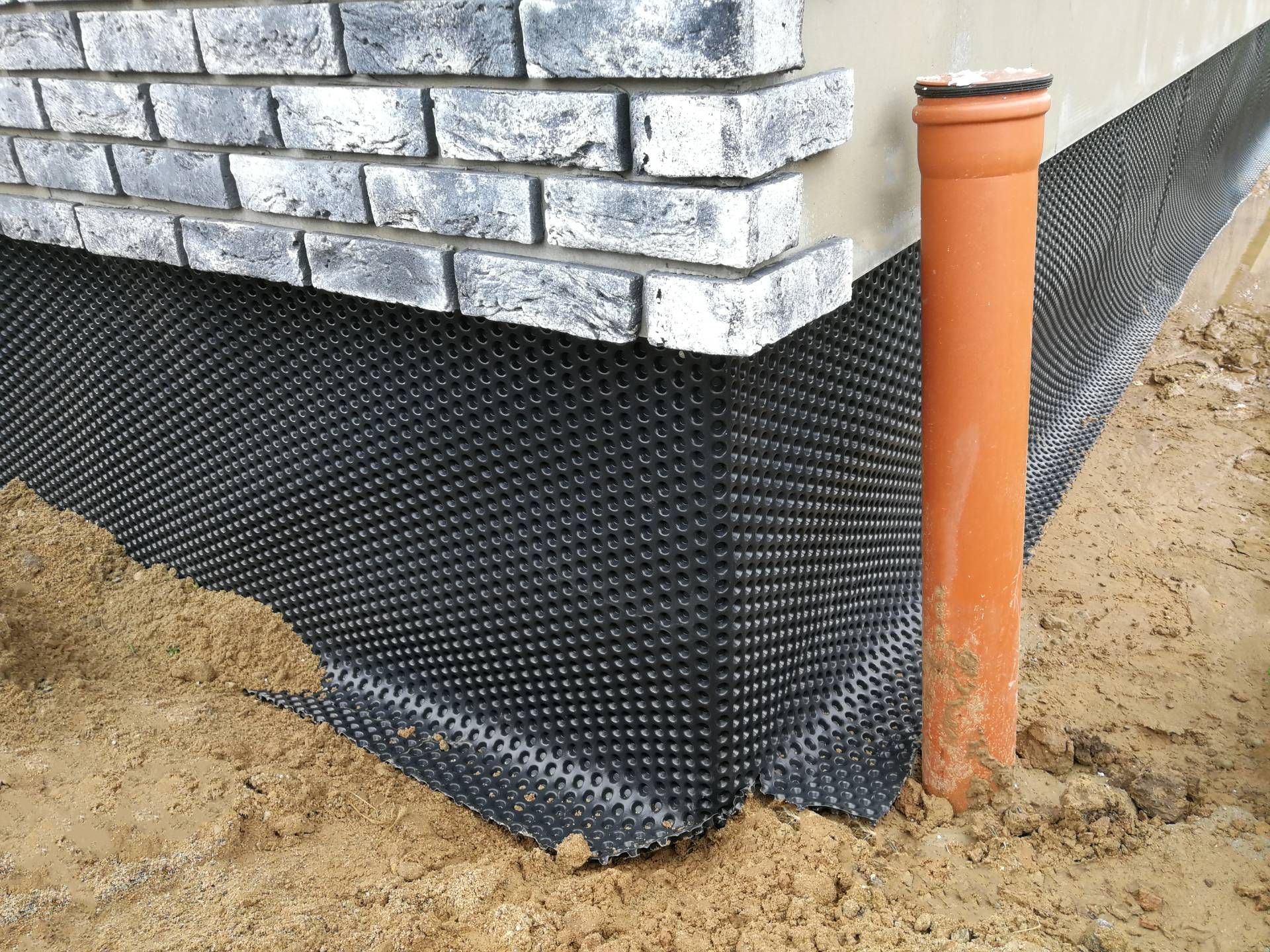 Black dimpled foundation wrap against a brick wall, next to a drainage pipe, set in soil.