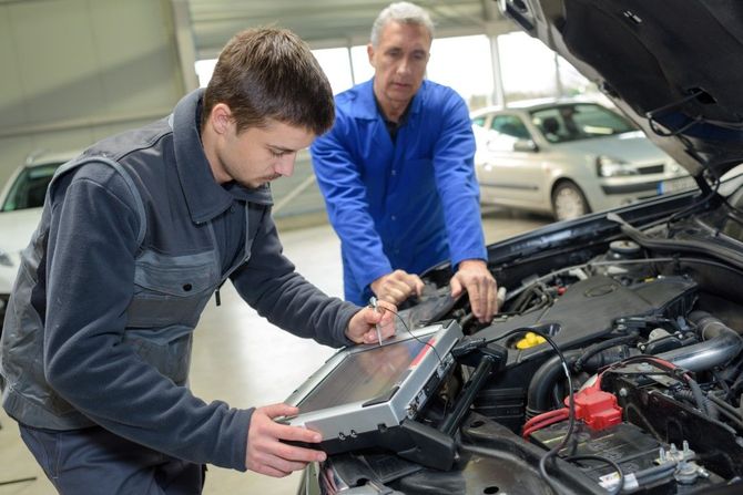Two mechanics in work uniforms examine an open car engine using a tablet computer in a garage.