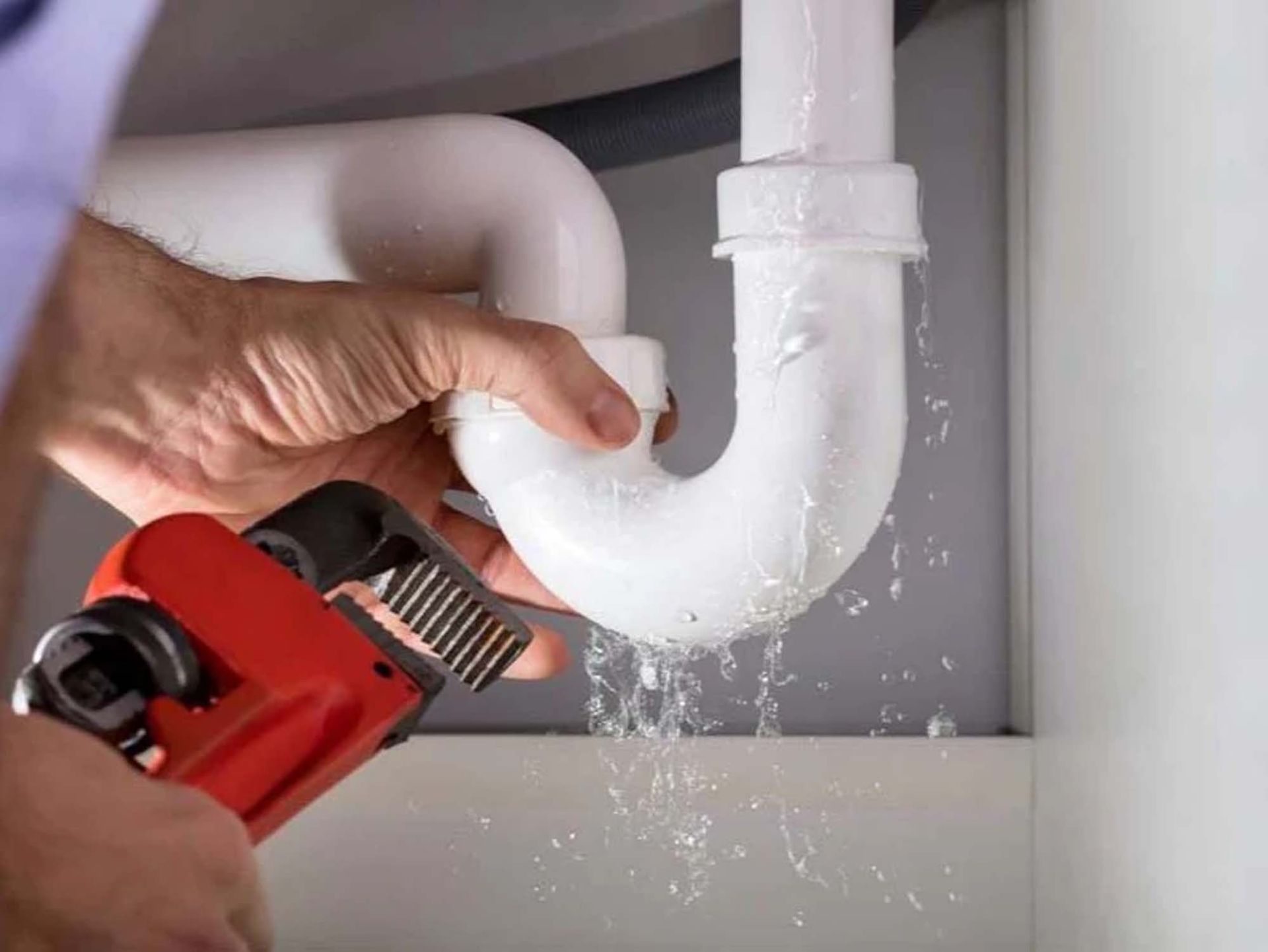 Plumber fixing a leaking white pipe under a sink, using a red wrench. Water is spraying.