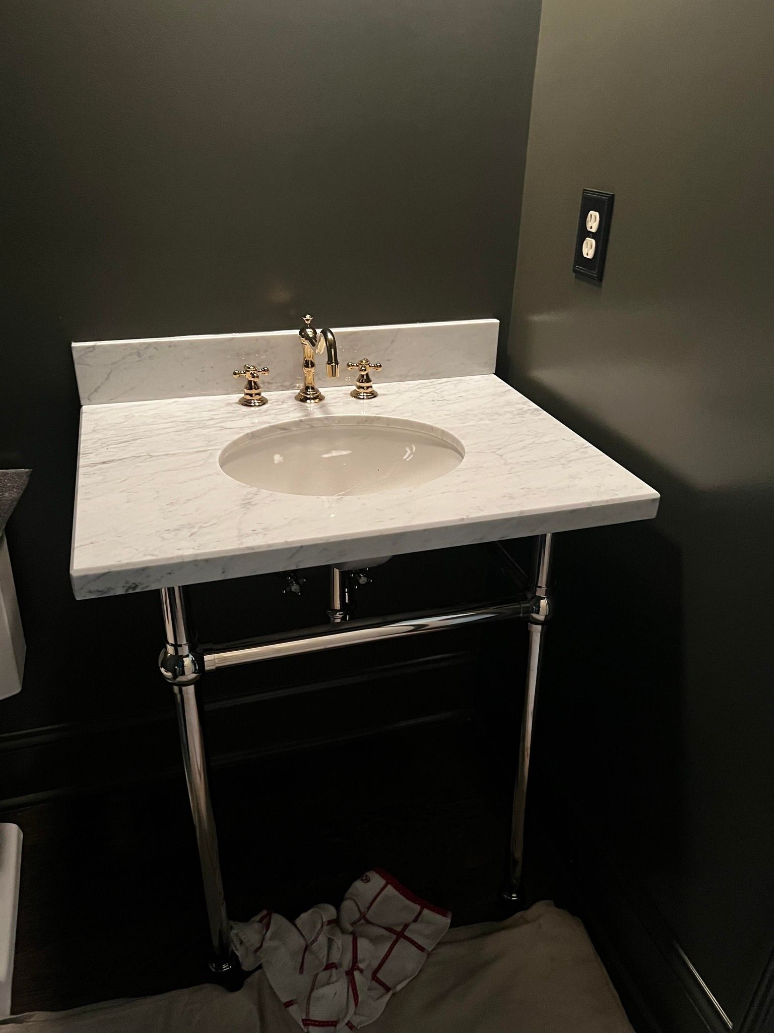 A marble countertop sink with gold fixtures and a metal stand in a dark-walled bathroom. A white towel lies on the floor.